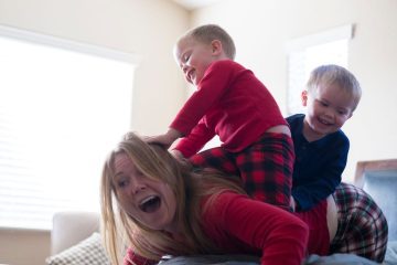 A mother is playfully engaging with her two young children on a bed. The children, dressed in matching red and blue plaid pajamas, are climbing over their mother, with big smiles on their faces as they have fun. The mother, laughing, is lying on her stomach while the boys joyfully sit on her back, creating an amusing and lively moment. The bright light from the window enhances the cheerful and energetic mood of the scene, reflecting a playful family interaction.